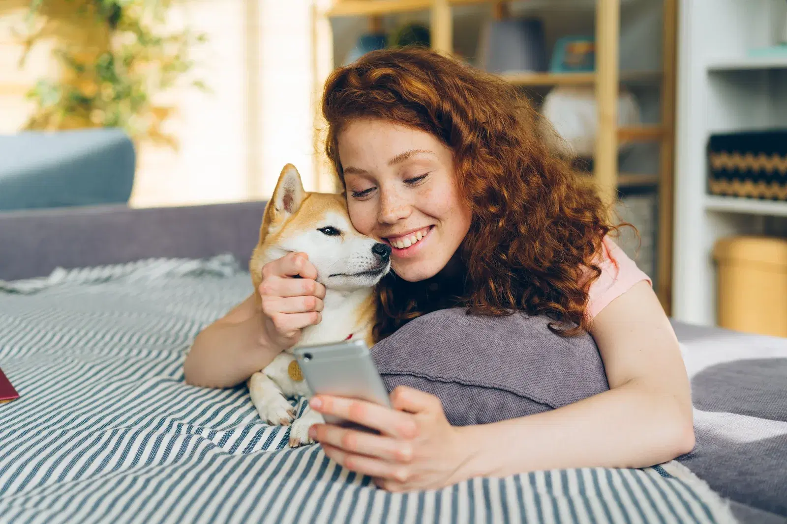 Woman lying on a bed with her Shiba Inu, smiling as she looks at her smartphone